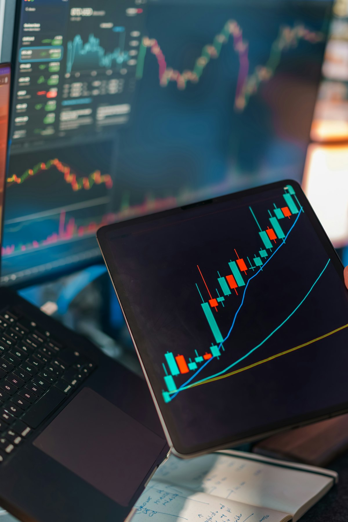 Tablet showing a candlestick chart beside monitors with price charts